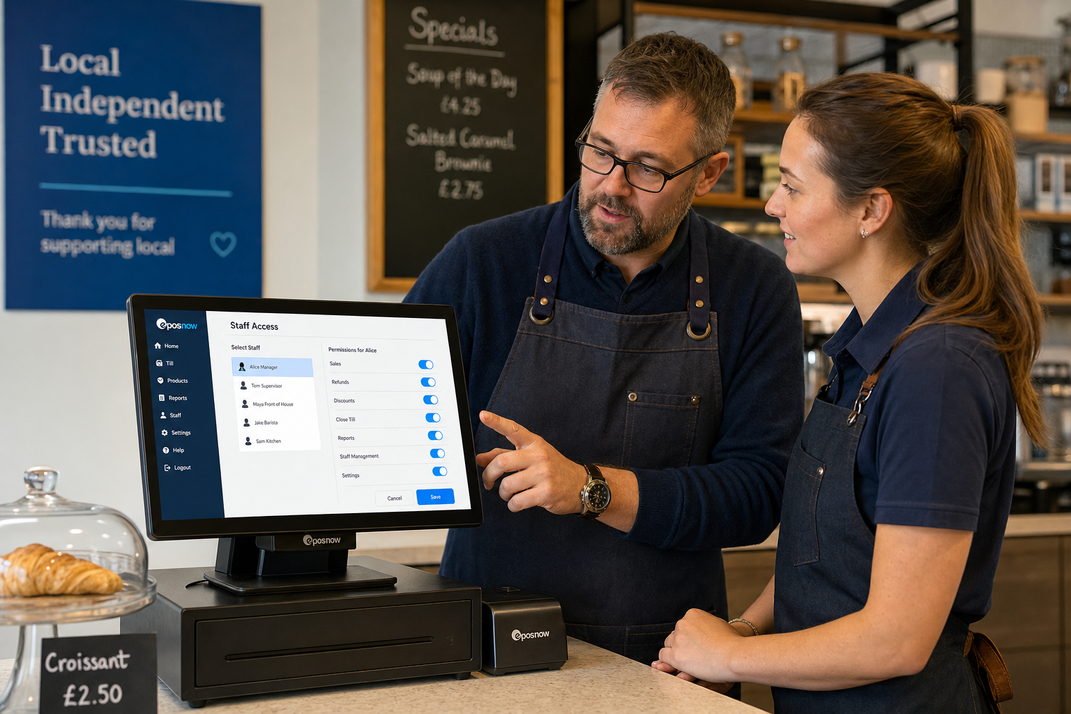 Why Staff Permissions Matter in Retail and Hospitality EPOS Systems - Photorealistic commercial photography of a modern UK independent business counter with a touchscreen EPOS terminal showing a clear staff login or user permissions screen, a supervisor and a front-of-house team member discussing access levels beside the till, with a café or small retail shop setting in the background, warm natural lighting, shallow depth of field, clean counter area, subtle blue brand accents on the interface and signage, premium realistic composition, friendly professional atmosphere, realistic POS hardware and a practical business setting that clearly communicates role-based access and day-to-day control.